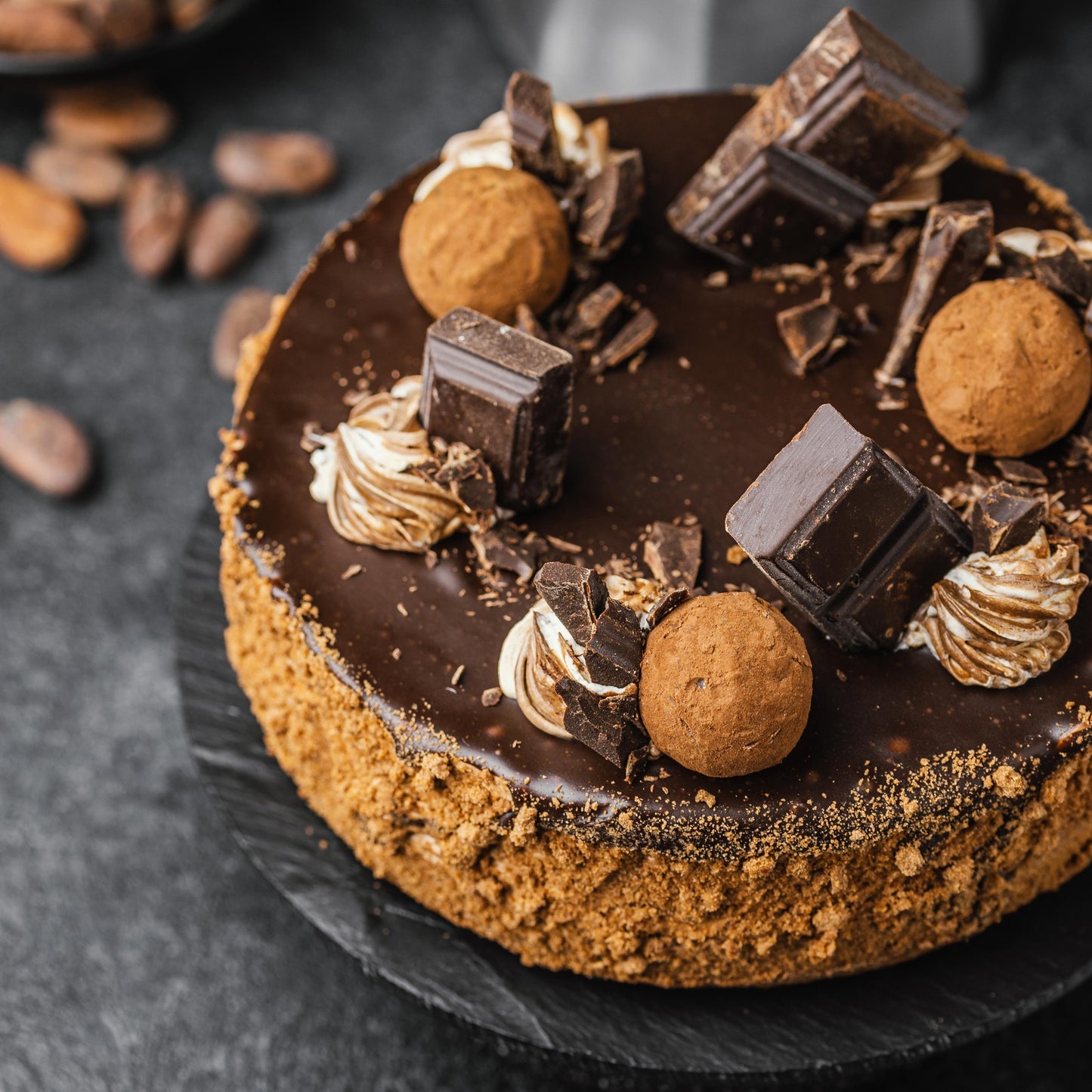 Angled top view of a dark chocolate cake with a caramelized biscuit crust, cocoa whipped cream dollops, chocolate bar chunks, and deep-fried choux balls.