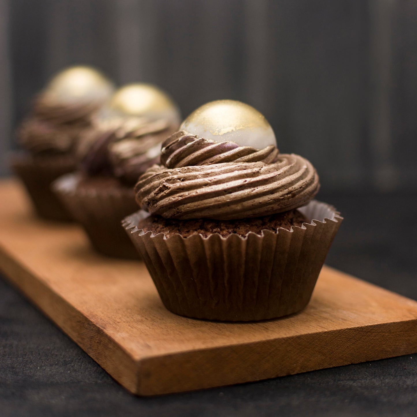 Front close-up view of three salted caramel cream-filled cupcakes with gold-dusted white chocolate caramel-filled balls, and chocolate buttercream swirls, on a wooden board.