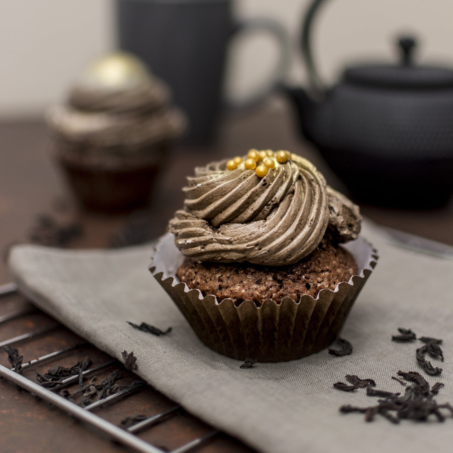 Angled front view of a salted caramel cream-filled cupcake with golden white chocolate caramel-filled mini-balls, and chocolate buttercream swirls, on a towel, with another cupcake, teapot, and mug used as decor in the background