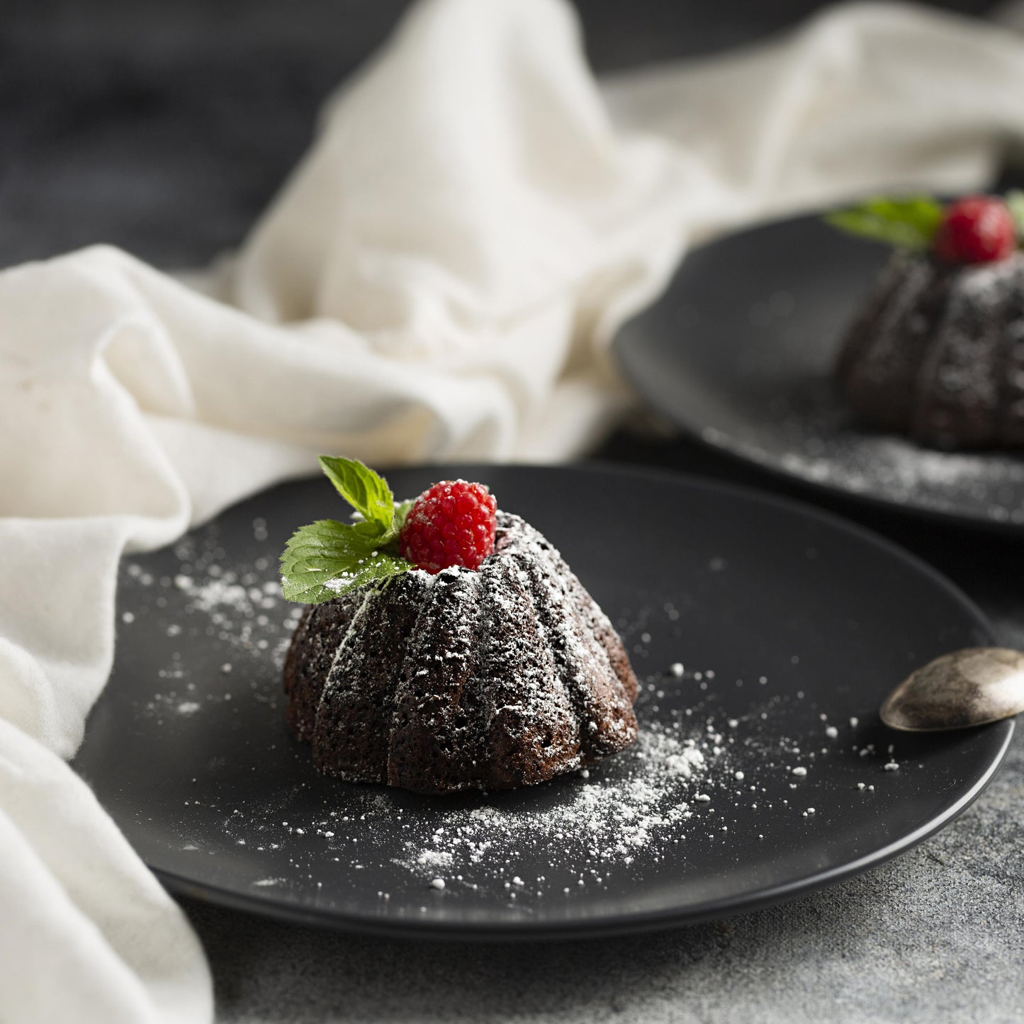 Angled front view of two chocolate mini cakes topped with confectioner sugar, fresh raspberries, and mint leaves, on black plates, with a teaspoon and cloth used as décor in the background.