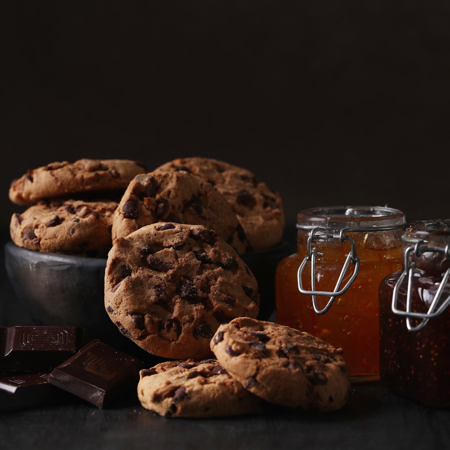 Front view of several chocolate chip cookies in front of a bowl with more chocolate chip cookies, with two square jars of jam, one with apricot and another one with raspberries, and chunks of chocolate in the background.