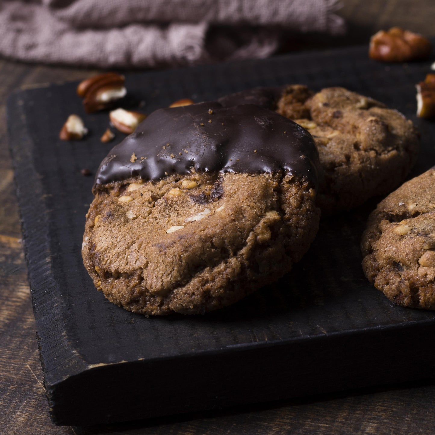 Angled top view of several chocolate-dipped double chocolate chip cookies on a black slate with walnuts and a light cloth in the background.