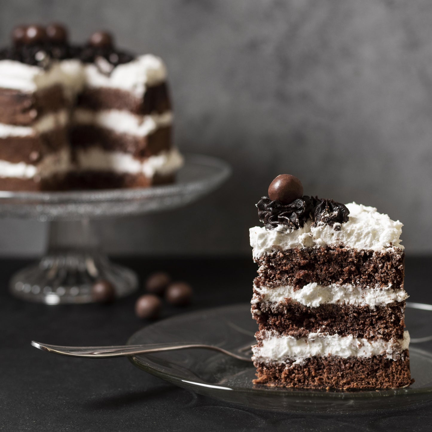 Front view of a cut moist chocolate cake on a glass cake stand and a moist chocolate cake slice on a glass plate, both layered with cream cheese frosting, decorated with cocoa buttercream dollops, and topped with milk chocolate-covered coffee beans.