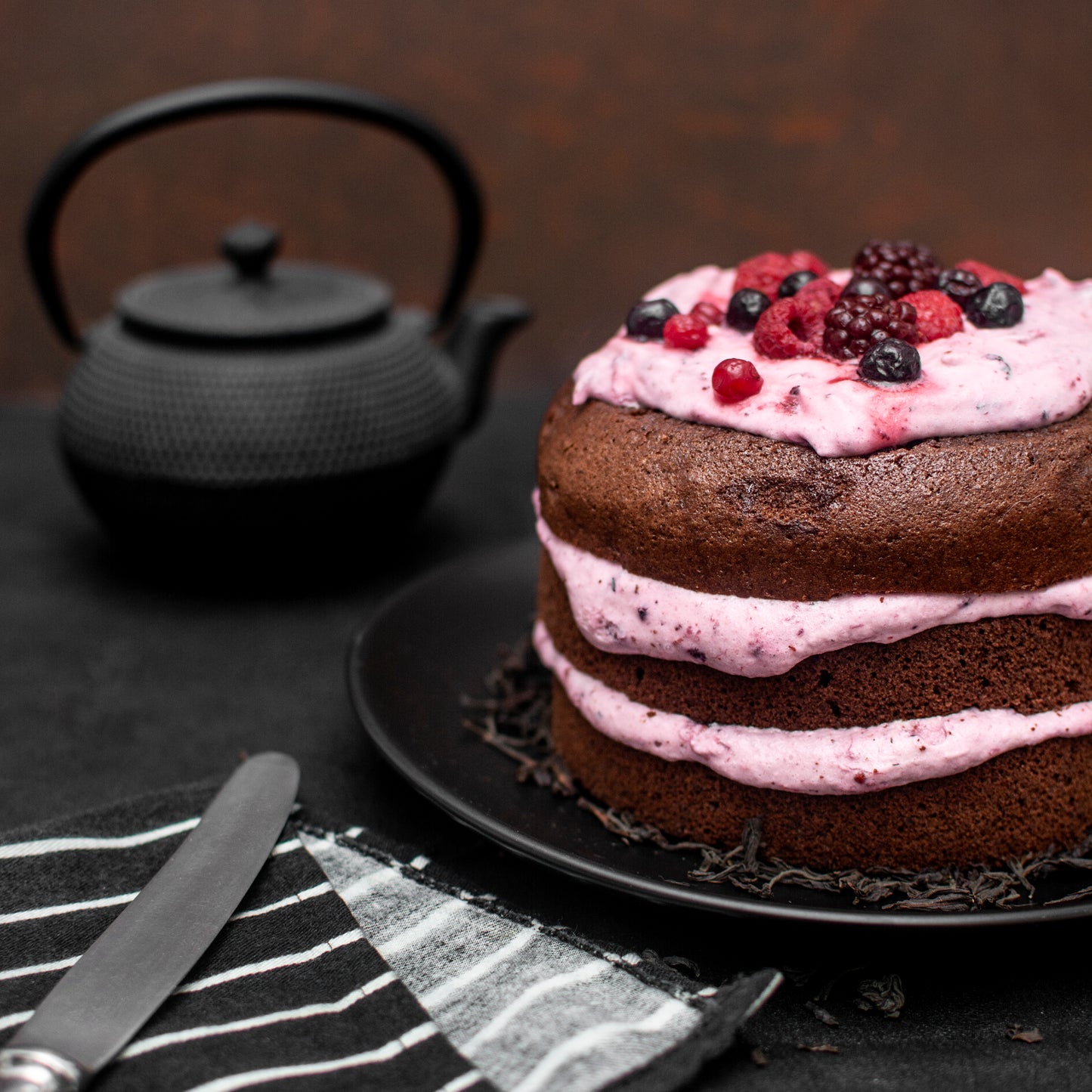 Front view cut close-up of a cocoa pound cake with layered berries yogurt foam, and fresh berries, on a black plate with a teapot and knife in the background.
