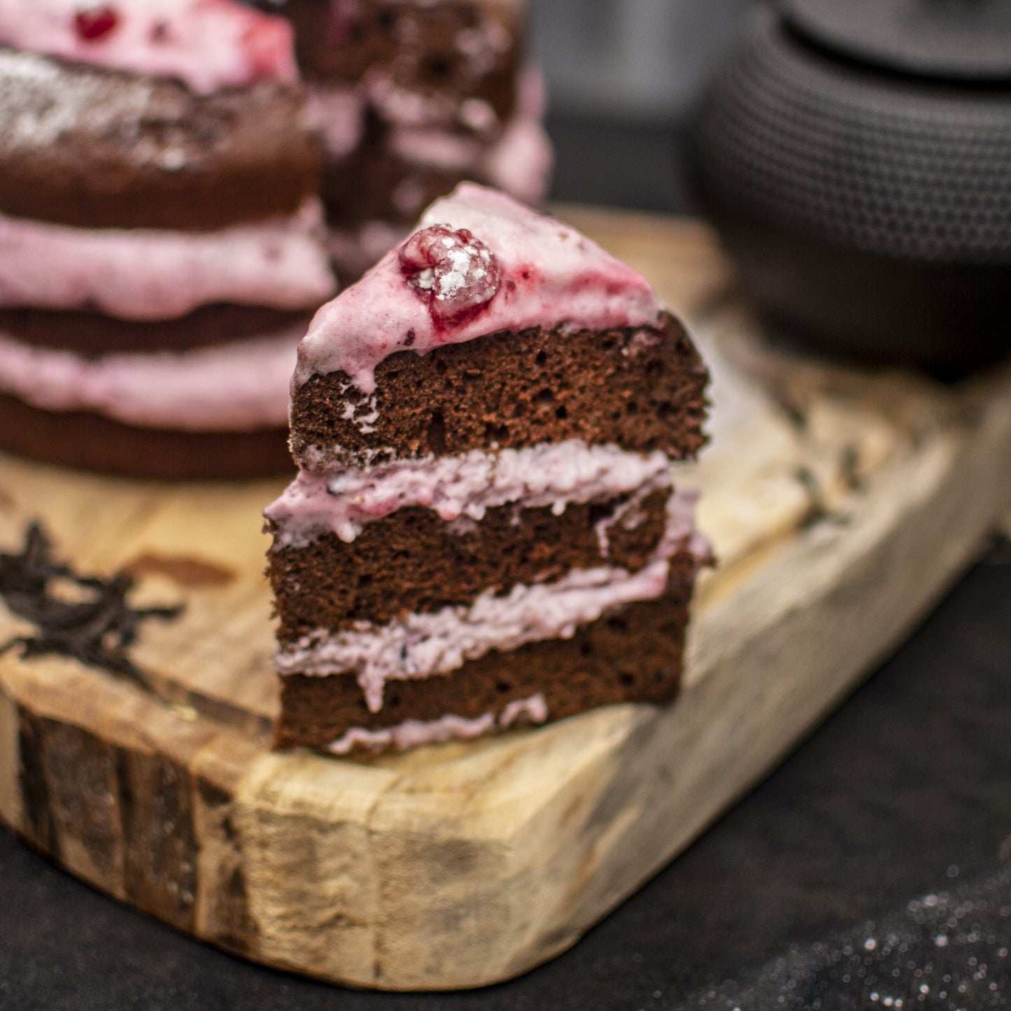 Front view of a cut cocoa pound cake and a cocoa pound cake slice, both layered with berries yogurt foam, and fresh berries, on a rustic wooden board with a teapot in the background.