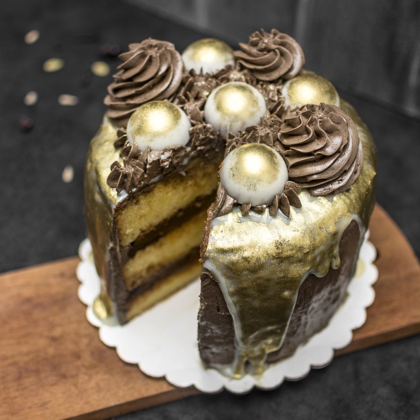 Angled top view of a cut salted caramel cream-filled sponge cake, covered with chocolate buttercream, drizzled with white chocolate ganache, edible liquid gold, decorated with gold-dusted white chocolate caramel-filled balls, and chocolate buttercream swirls, on a wooden board.
