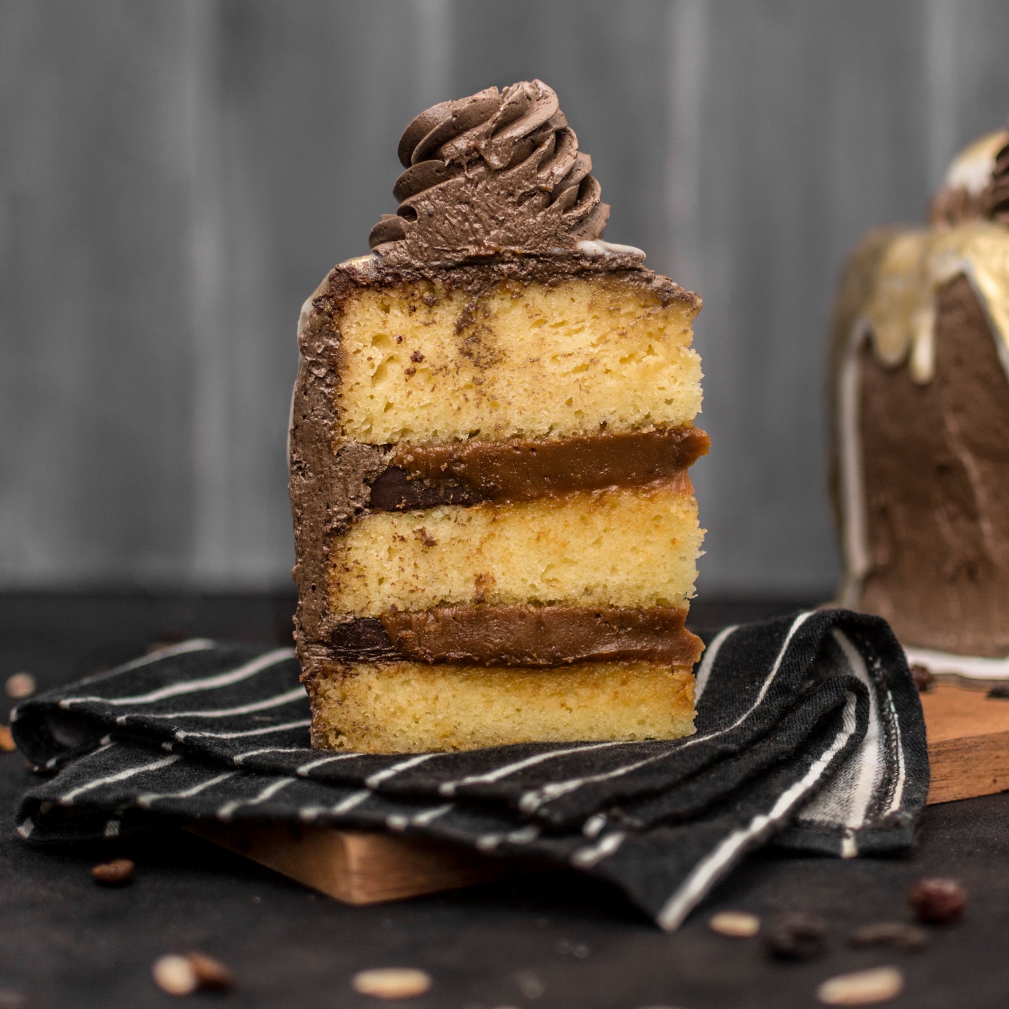 Front view of a salted caramel cream-filled sponge cake slice, covered with chocolate buttercream, drizzled with white chocolate ganache, edible liquid gold, decorated with gold-dusted white chocolate caramel-filled balls, and chocolate buttercream swirls, on a dark cloth placed on wooden board.