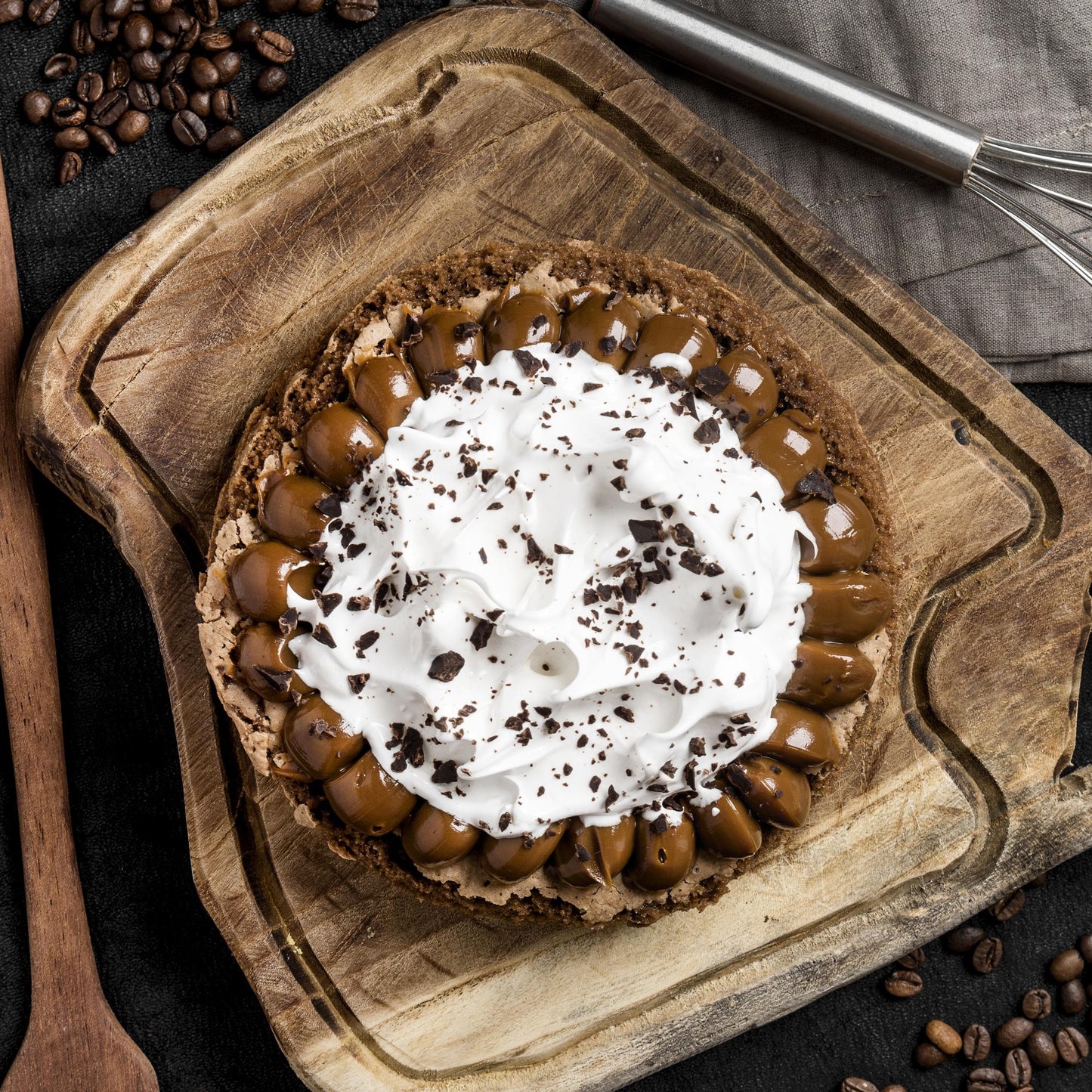 Top view of a brownie cake crowned with Italian meringue and salted caramel dollops, on a rustic wooden board with a cloth, mug with coffee, whisk, wooden spoon, and coffee beans in the background.