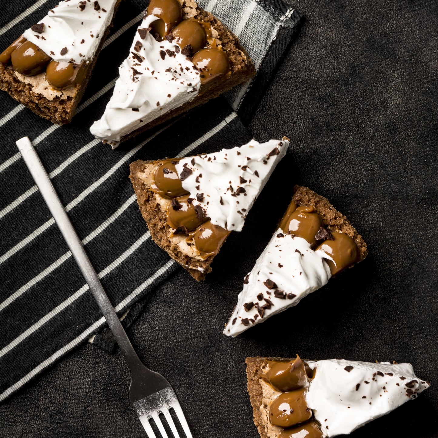 Top view of 5 brownie cake slices crowned with Italian meringue and salted caramel dollops, on a cloth with a fork in the background.
