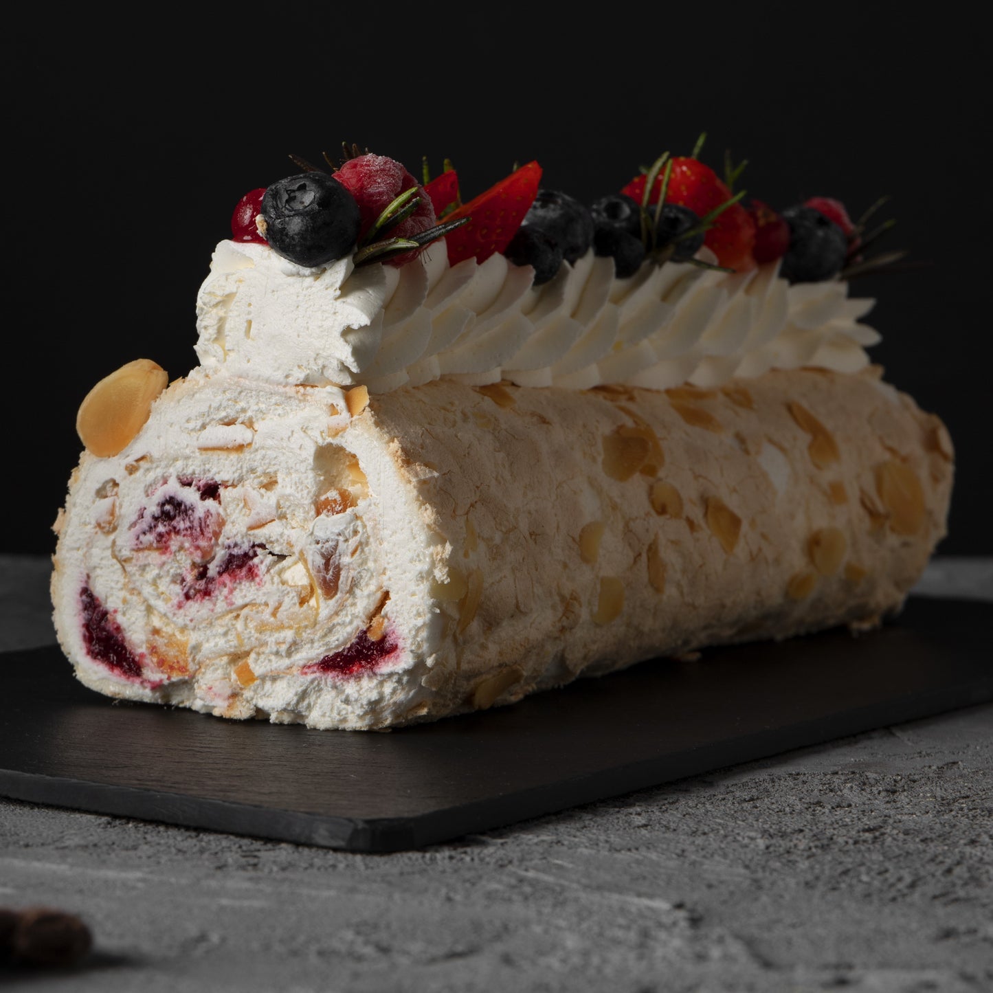 Front view of a tropical rolled cake with whipped cream swirls, açai compote filling, fresh berries décor and rosemary leaves, on a black slate board.