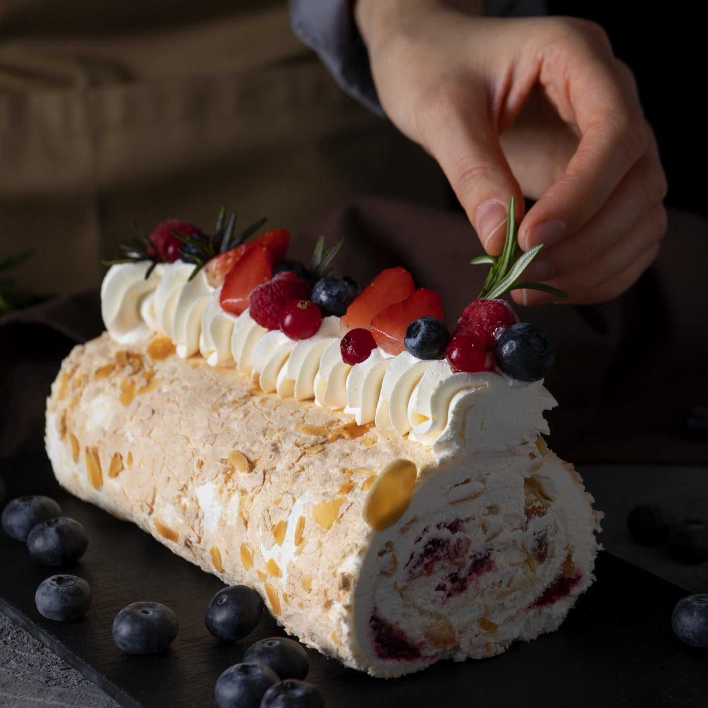 Angled front view of a tropical rolled cake with whipped cream swirls, açai compote filling, fresh berries décor and rosemary leaves, on a black slate board with scattered blueberries and a hand decorating the cake in the background.
