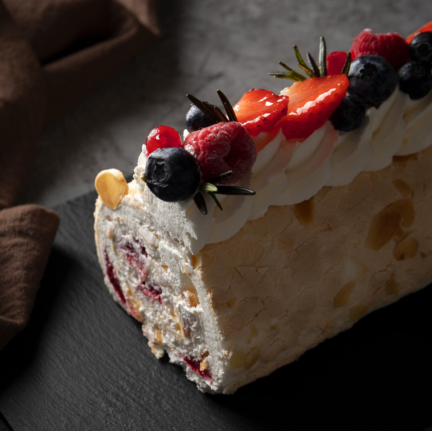 Angled cut top view of a tropical rolled cake with whipped cream swirls, açai compote filling, fresh berries décor and rosemary leaves, on a black slate board with a cloth in the background.