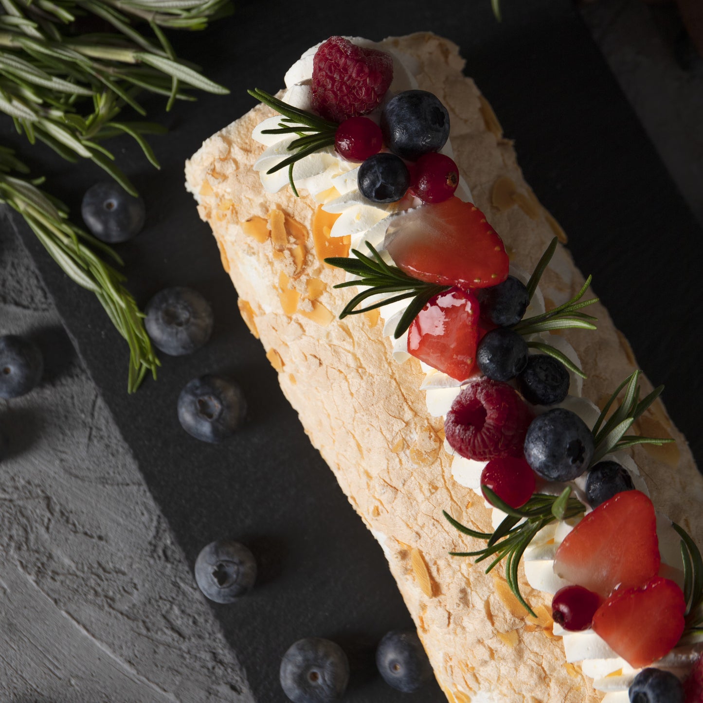 Top view of a tropical rolled cake with whipped cream swirls, açai compote filling, fresh berries décor and rosemary leaves, on a black slate board with a cloth, rosemary sprigs, and scattered blueberries in the background.
