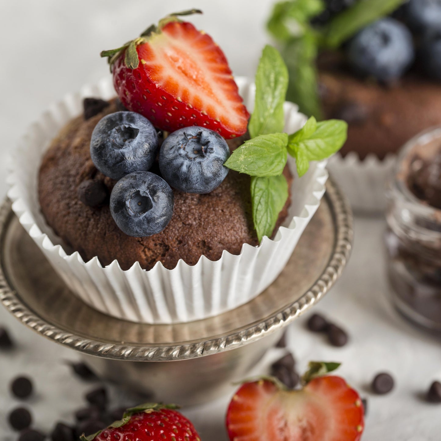 Angled top view of a chocolate chip cupcake topped with fresh forest fruits and mint leaves, on an elevated metal stand, scattered forest fruits chocolate chips, a jar of chocolate spread, and another cupcake used as décor in the background.