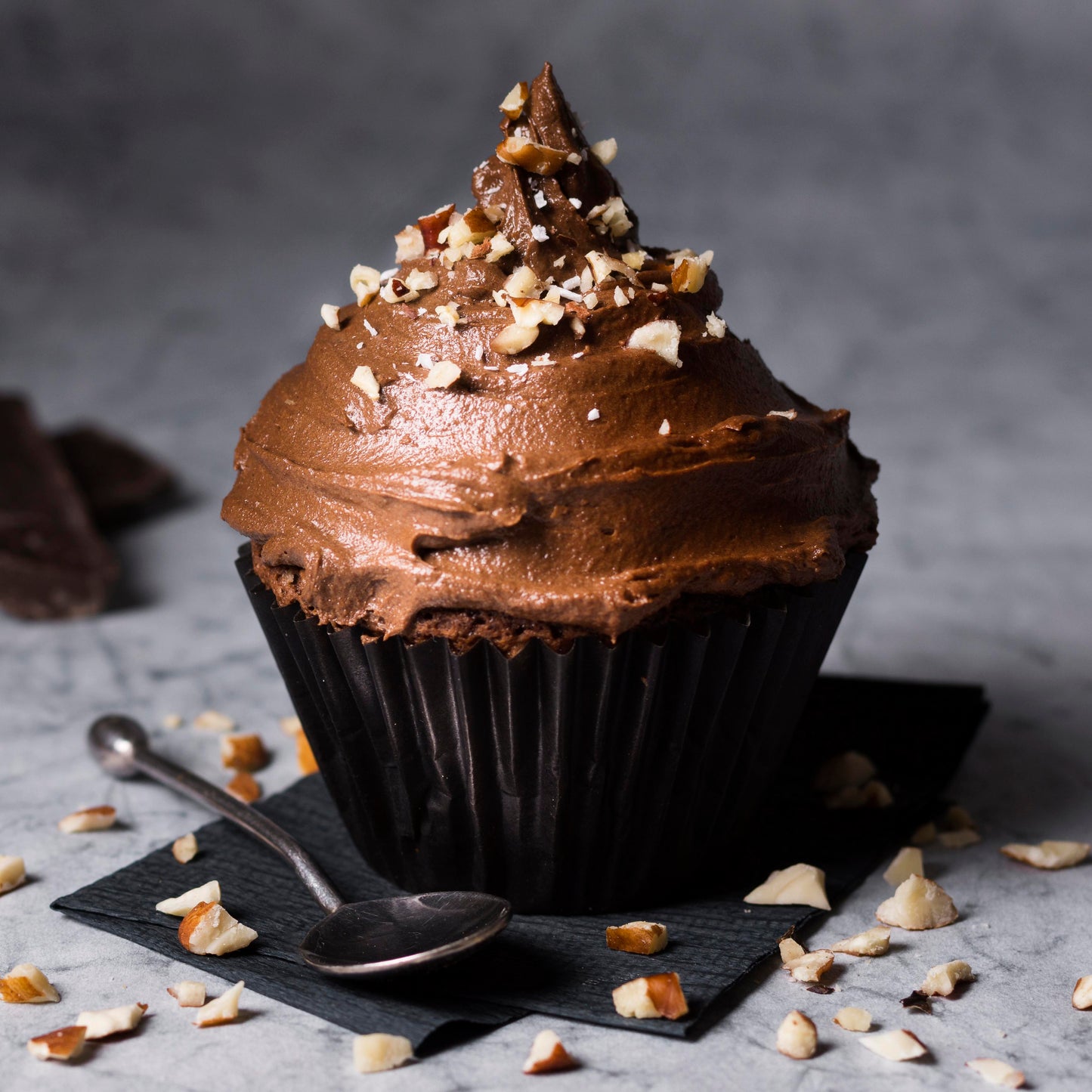Front close-up view of a chocolate giant cupcake decorated with a mix of hazelnut-chocolate buttercream and topped with roasted hazelnut crisps, on a black cloth with a teaspoon, scattered chocolate chips, and hazelnut crisps used as décor in the background.
