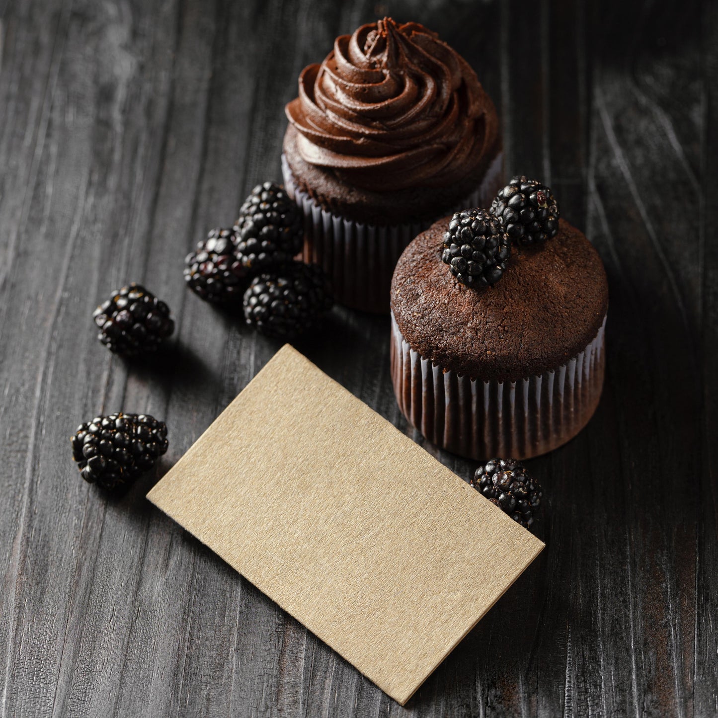 Angled top view of a kraft-colored paper card and two tall chocolate ramekin-shaped cupcakes, one decorated with chocolate buttercream and the other topped with fresh blackberries.