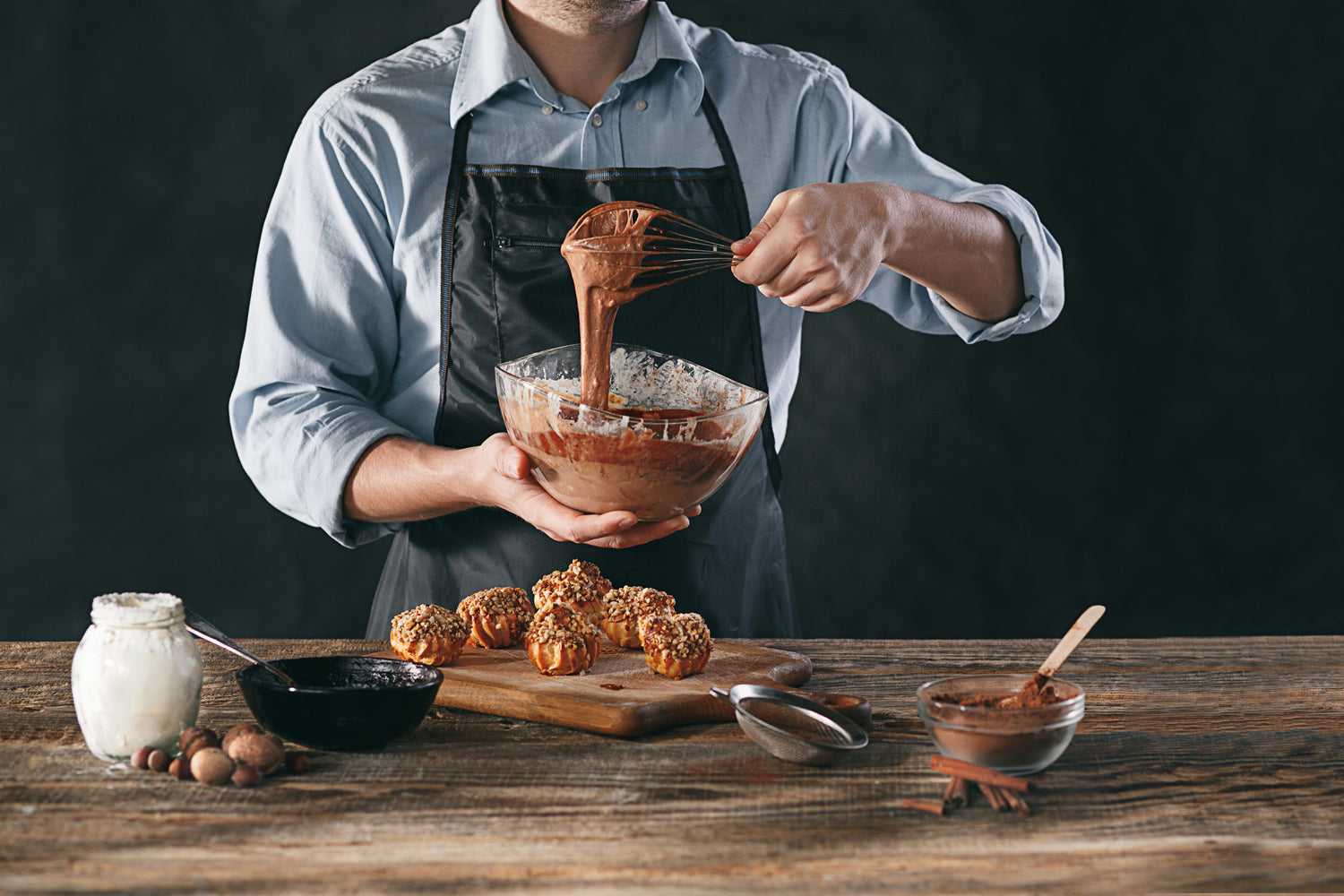 Front view of a chef showing a transparent bowl of chocolate filling with choux balls on a cutting board and baking ingredients laying on a wooden table.