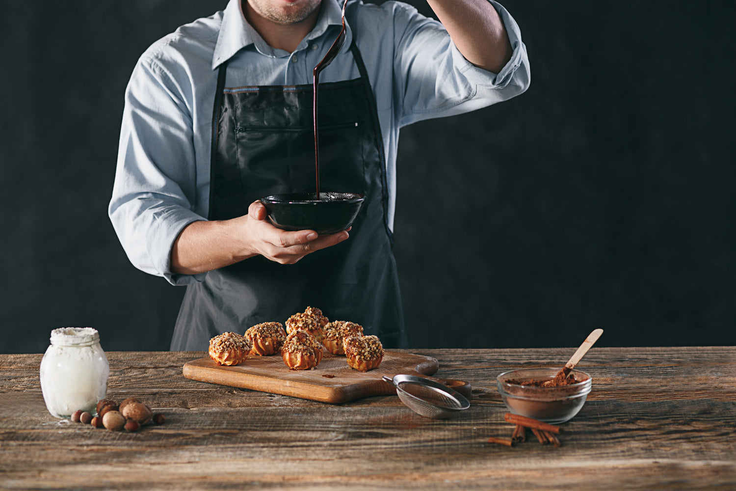 Front view of a chef showing a black bowl of dripping chocolate sauce with choux balls on a cutting board and baking ingredients laying on a wooden table.