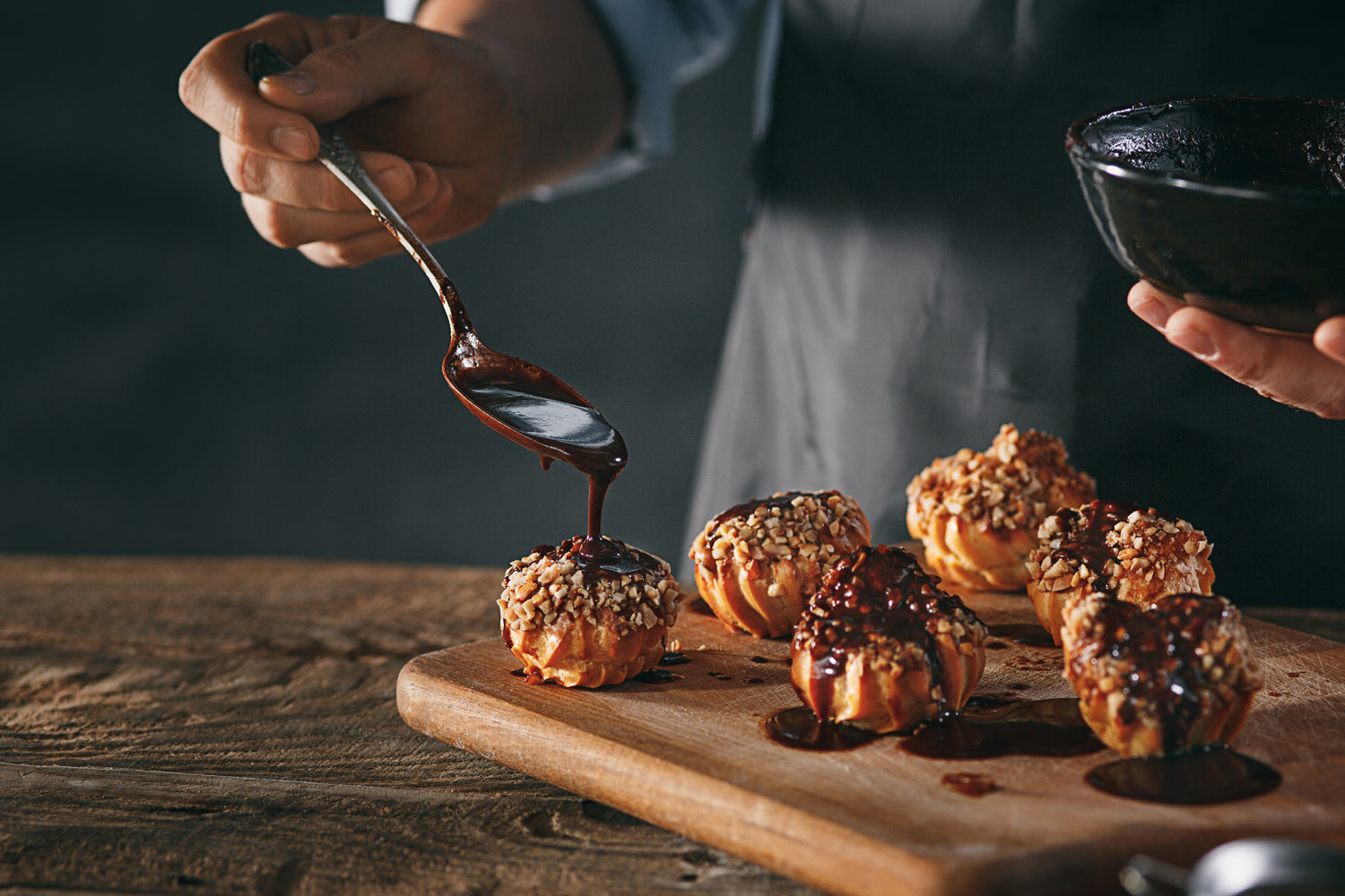 Angled close-up front view of a chef's hand drizzling chocolate sauce over choux balls that are on a cutting board laying around a wooden table.