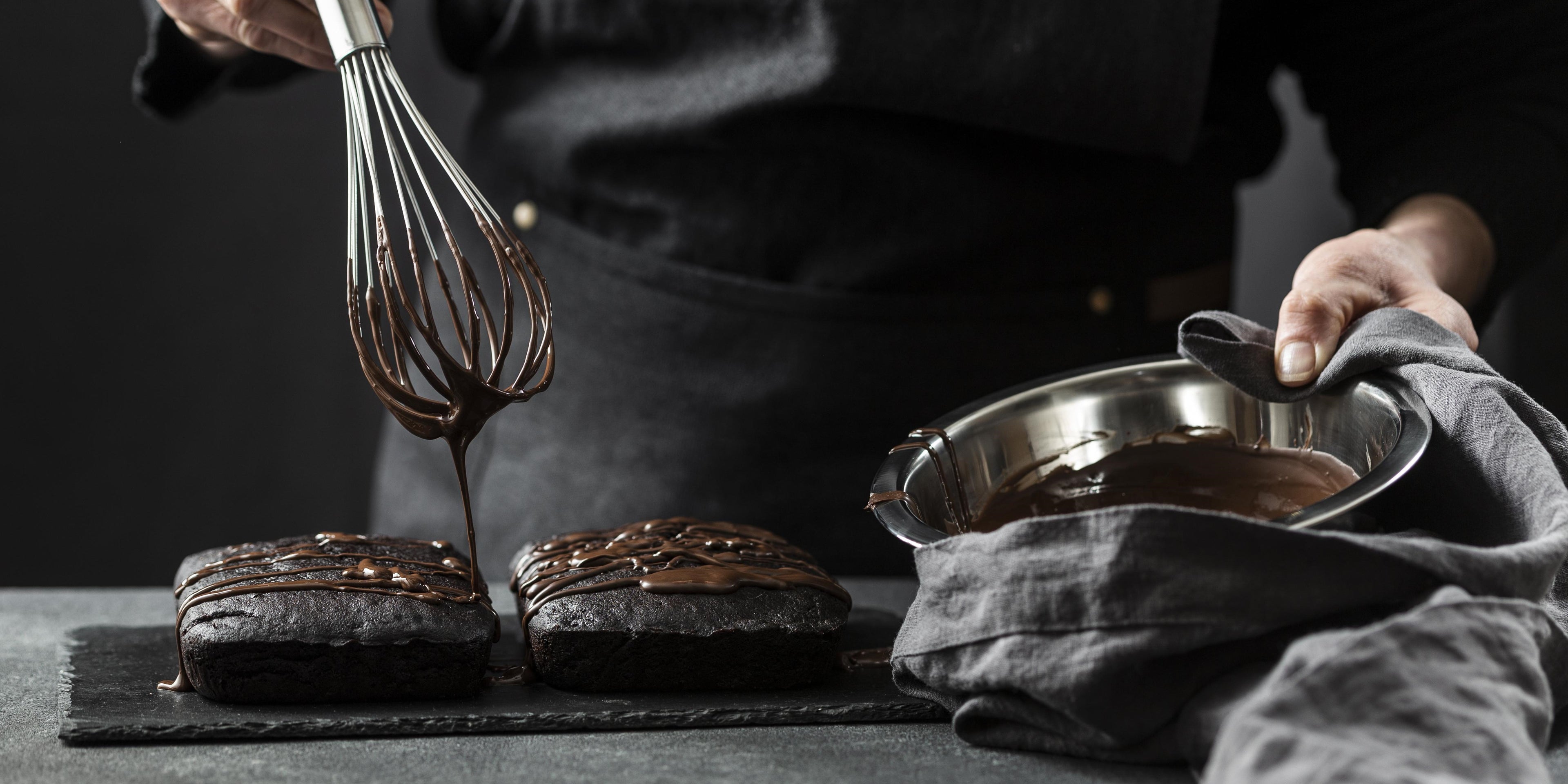 Front view of a person drizzling chocolate sauce over two brownie cakes on a black slate plate, with a whisk and holding a stainless steel bowl with a cloth.