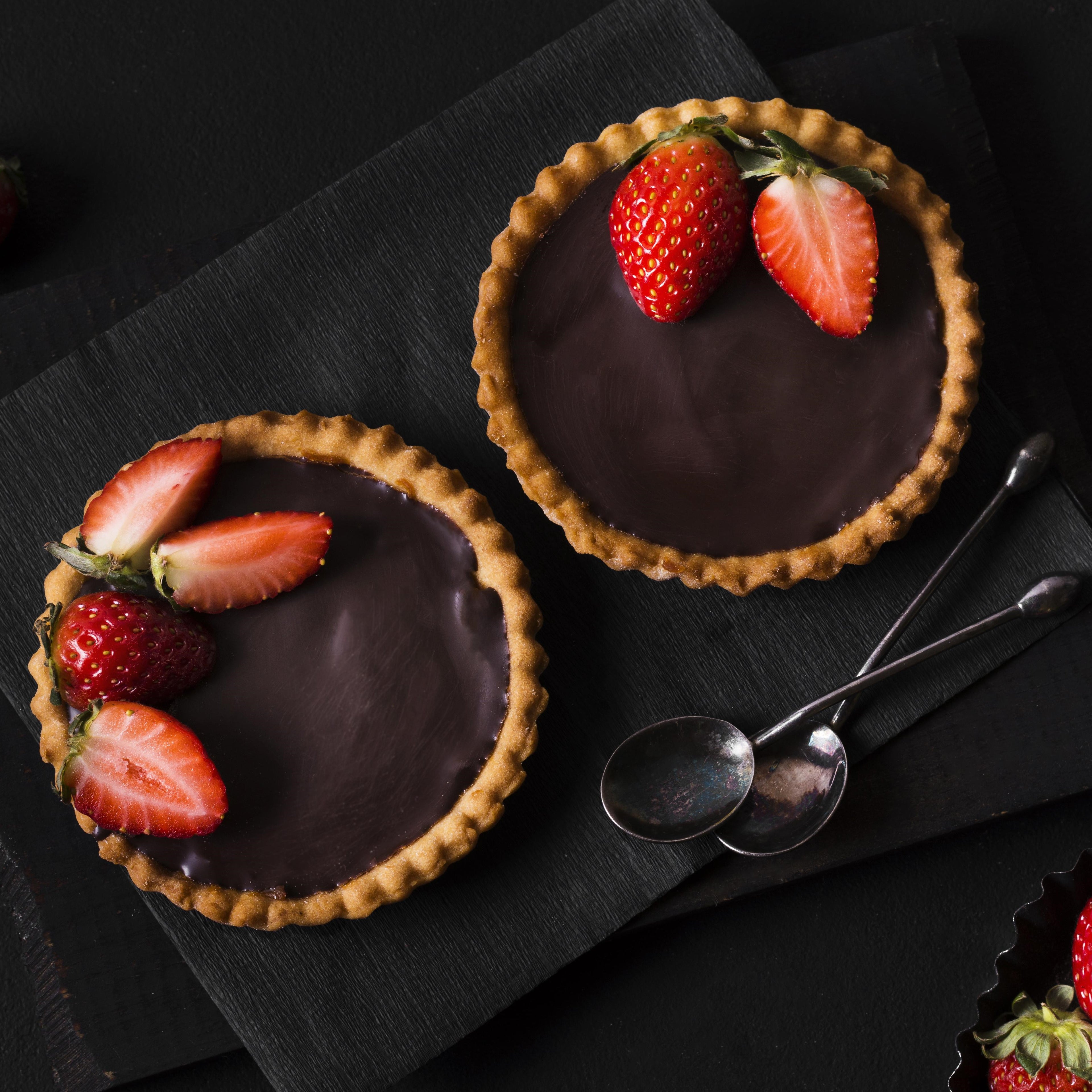 Top view of two mini chocolate tarts with halved strawberries on a dark slate plate with two teaspoons next to them.