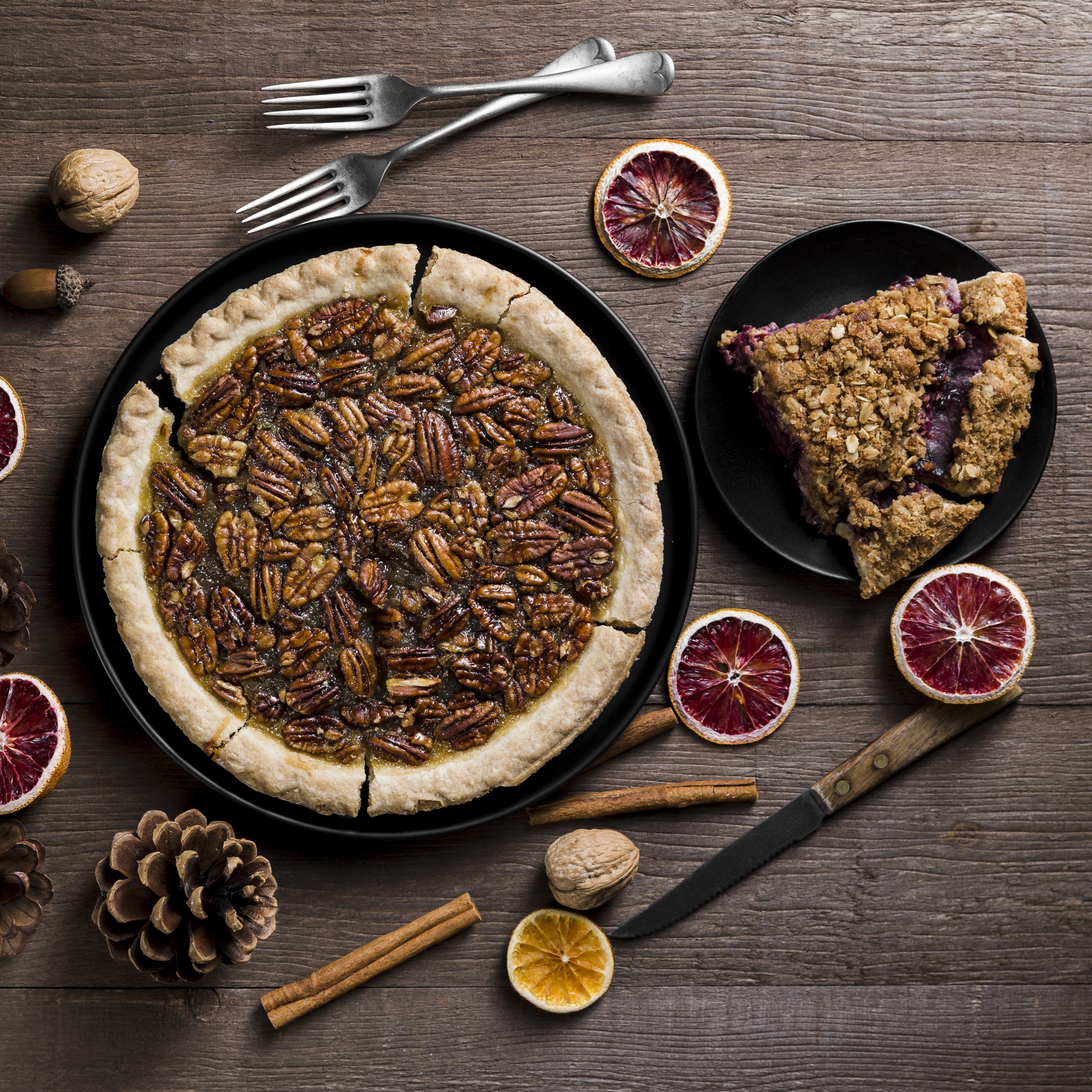 Top view of a pecan pie on a black plate with a slice of an unknown pie, decorated with dried blood orange slices.