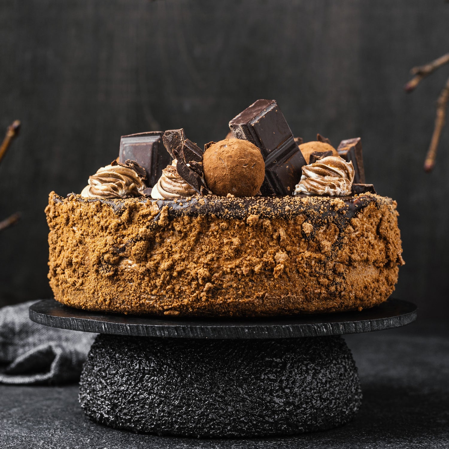 Front view of a dark chocolate cake with a caramelized biscuit crust, cocoa whipped cream dollops, chocolate bar chunks, and deep-fried choux balls.