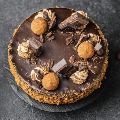 Top view of a dark chocolate cake with a caramelized biscuit crust, cocoa whipped cream dollops, chocolate bar chunks, and deep-fried choux balls.