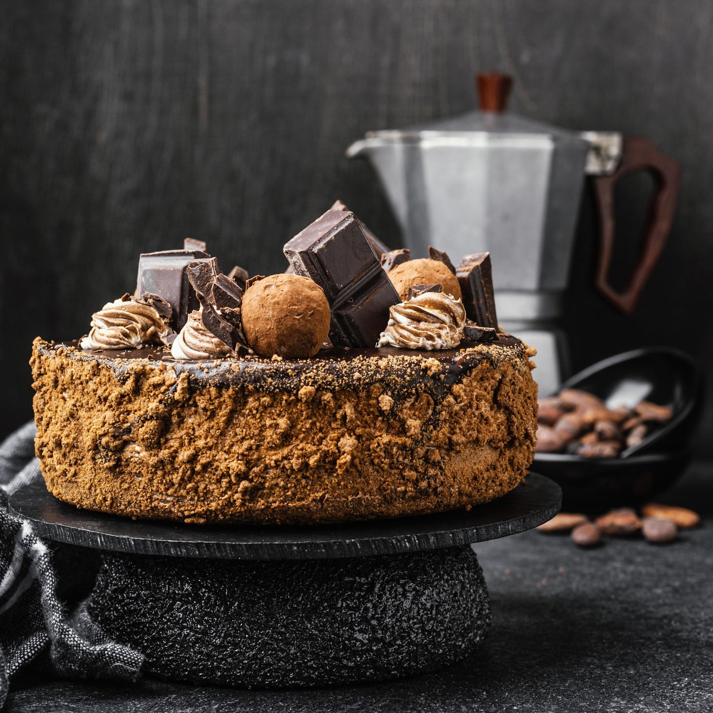 Angled front view of a dark chocolate cake with a caramelized biscuit crust, cocoa whipped cream dollops, chocolate bar chunks, and deep-fried choux balls.