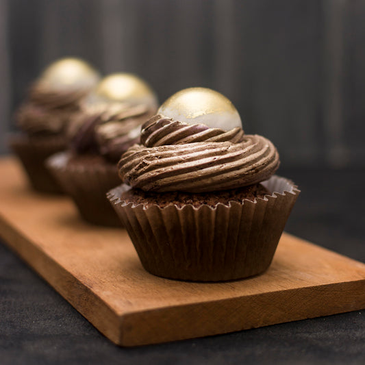Front close-up view of three salted caramel cream-filled cupcakes with gold-dusted white chocolate caramel-filled balls, and chocolate buttercream swirls, on a wooden board.