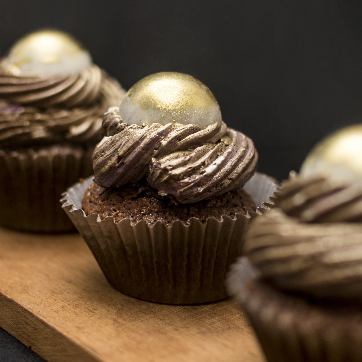 Front view of three salted caramel cream-filled cupcakes with gold-dusted white chocolate caramel-filled balls, and chocolate buttercream swirls, on a wooden board.
