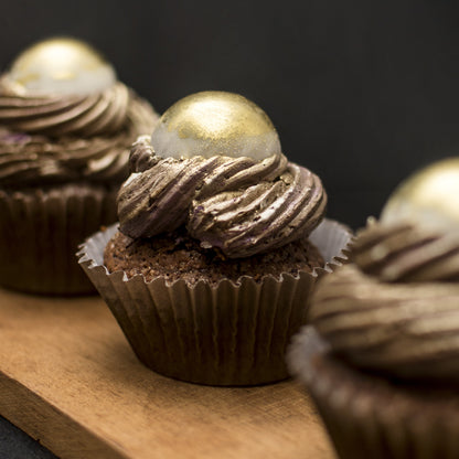Front view of three salted caramel cream-filled cupcakes with gold-dusted white chocolate caramel-filled balls, and chocolate buttercream swirls, on a wooden board.