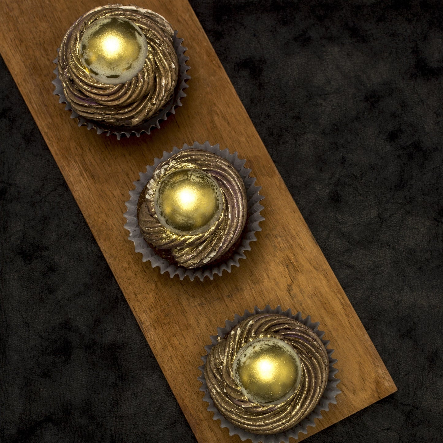 Top view of three salted caramel cream-filled cupcakes with gold-dusted white chocolate caramel-filled balls, and chocolate buttercream swirls, on a wooden board.