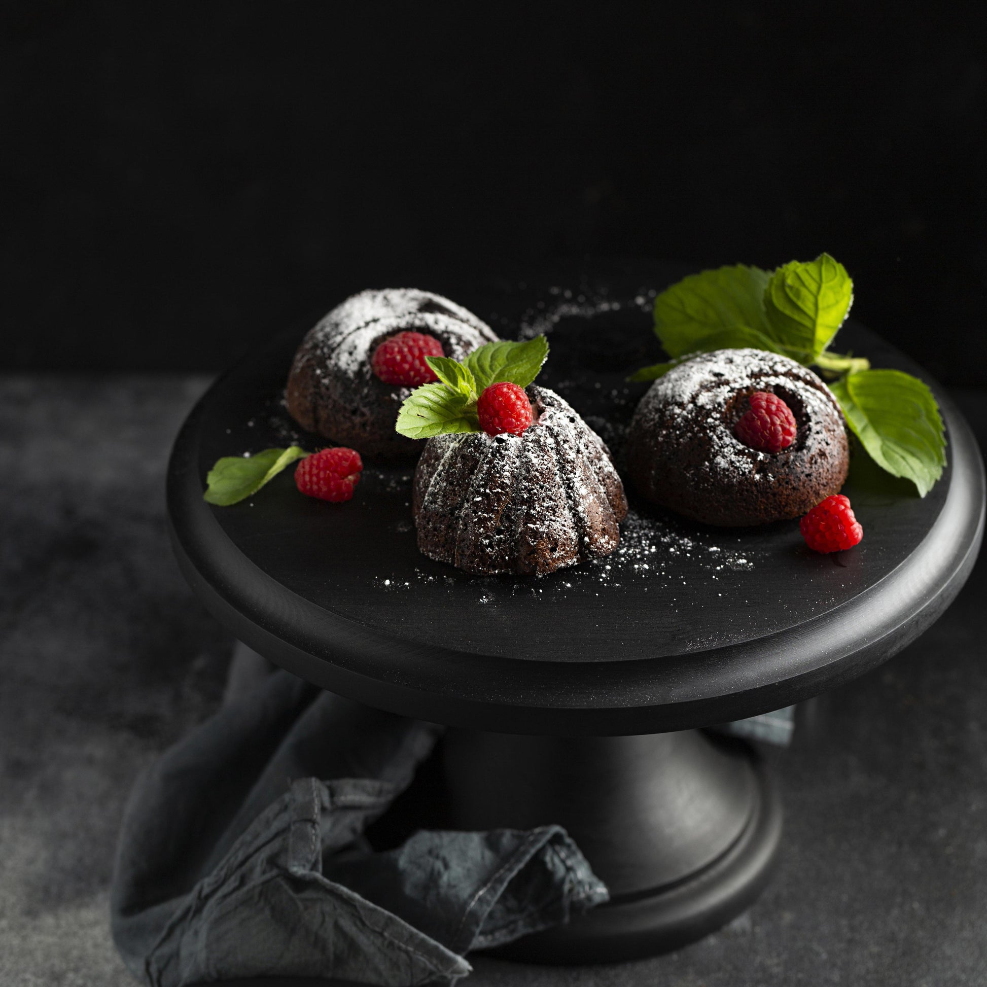 Angled top view of several chocolate mini cakes topped with confectioner sugar, fresh raspberries, and mint leaves, on a black wooden cake stand.