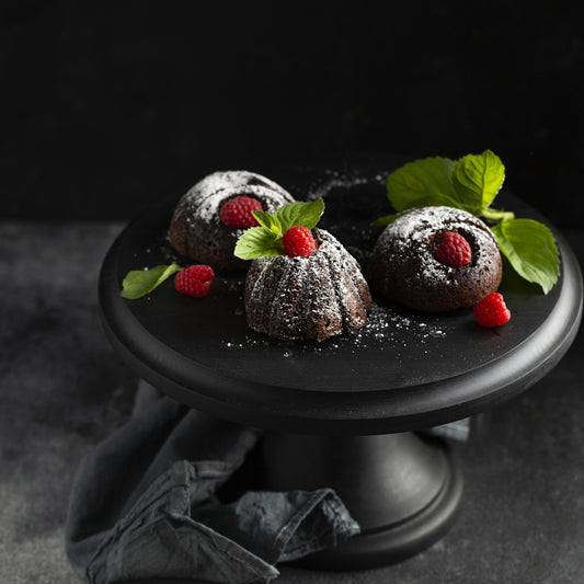 Angled top view of several chocolate mini cakes topped with confectioner sugar, fresh raspberries, and mint leaves, on a black wooden cake stand.