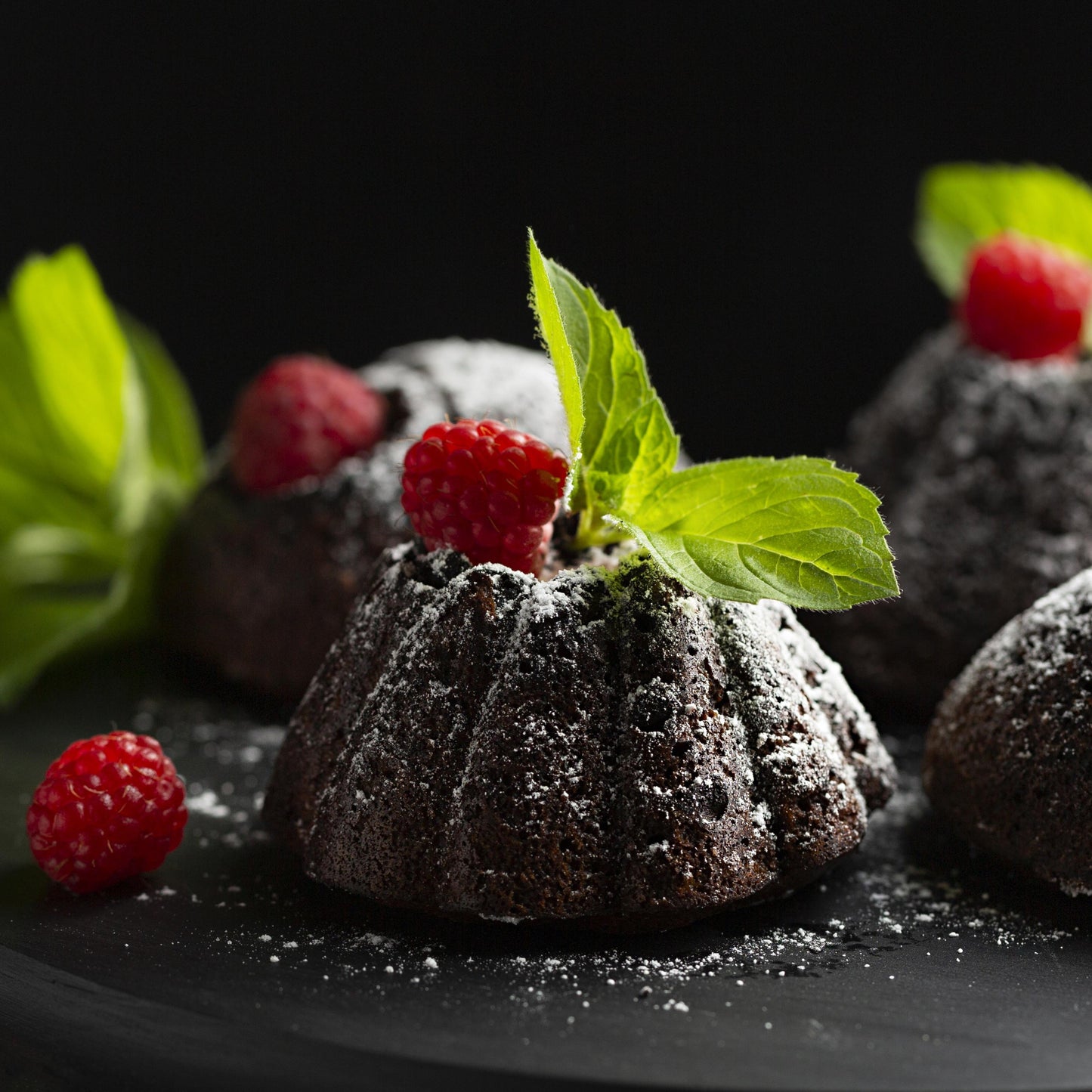 Front close-up view of several chocolate mini cakes topped with confectioner sugar, fresh raspberries, and mint leaves, on a black wooden board.