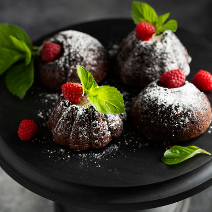 Angled top close-up view of several chocolate mini cakes topped with confectioner sugar, fresh raspberries, and mint leaves, on a black wooden cake stand.