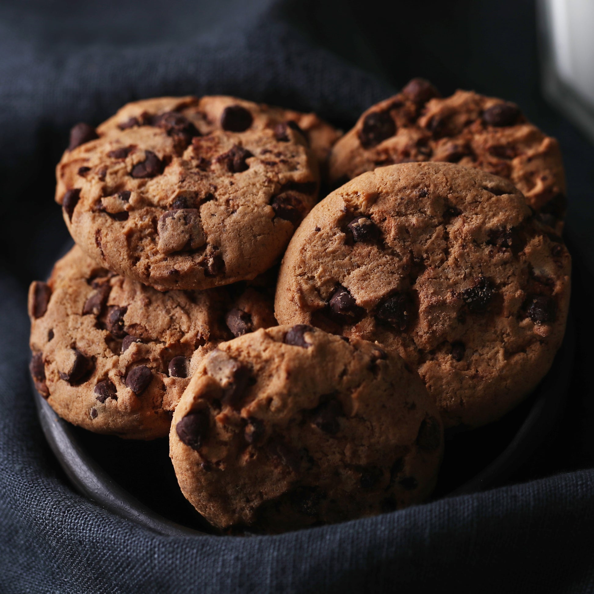 Angled top view of several chocolate chip cookies in a black plate with a dark cloth in the background.
