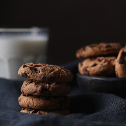Front view of a stack of several chocolate chip cookies placed on a dark cloth, with a bowl of more cookies and a glass of milk in the background.