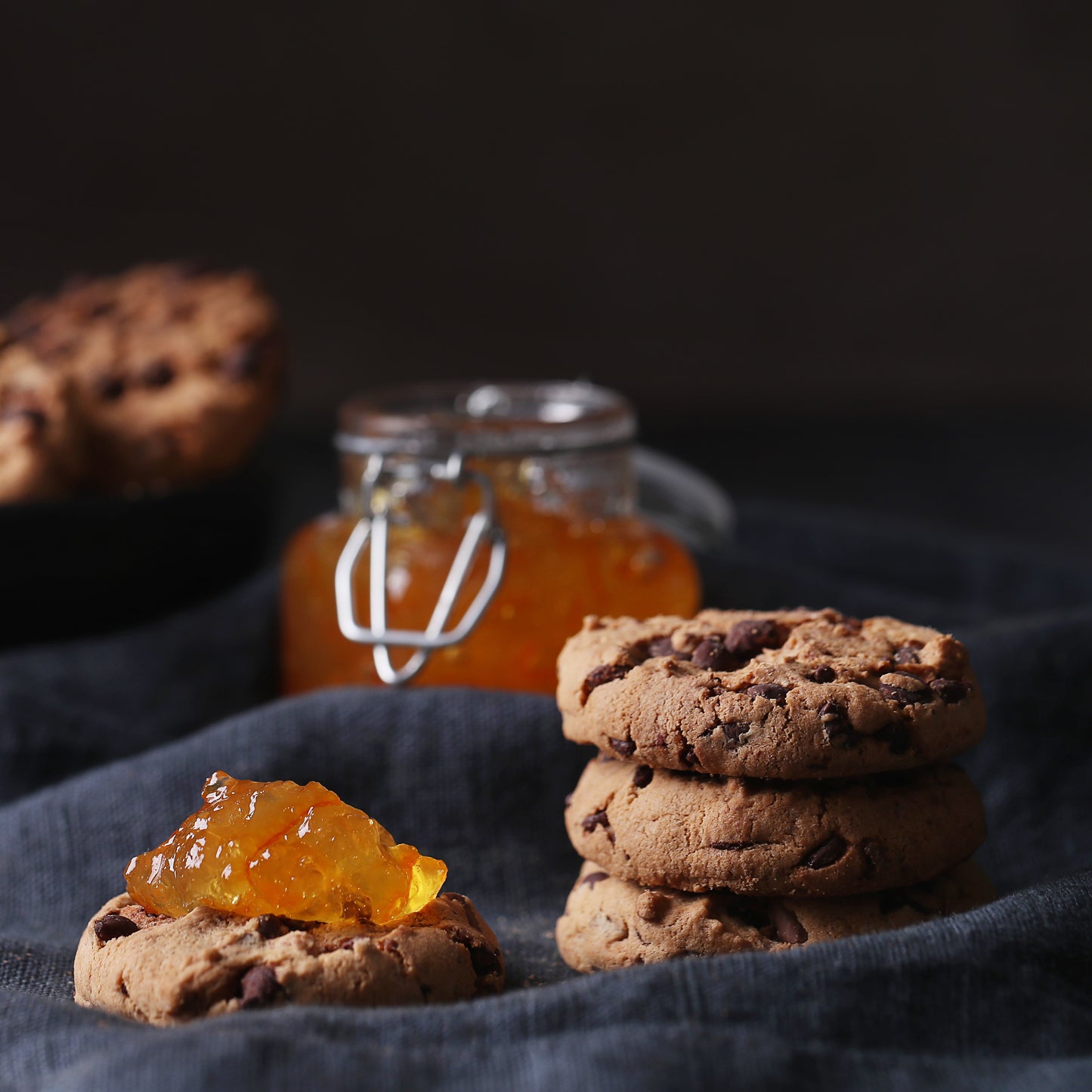 Front view of of a stack of several chocolate chip cookies and a cookie topped with apricot jam on a dark cloth, with a square jar of apricot jam in the background.