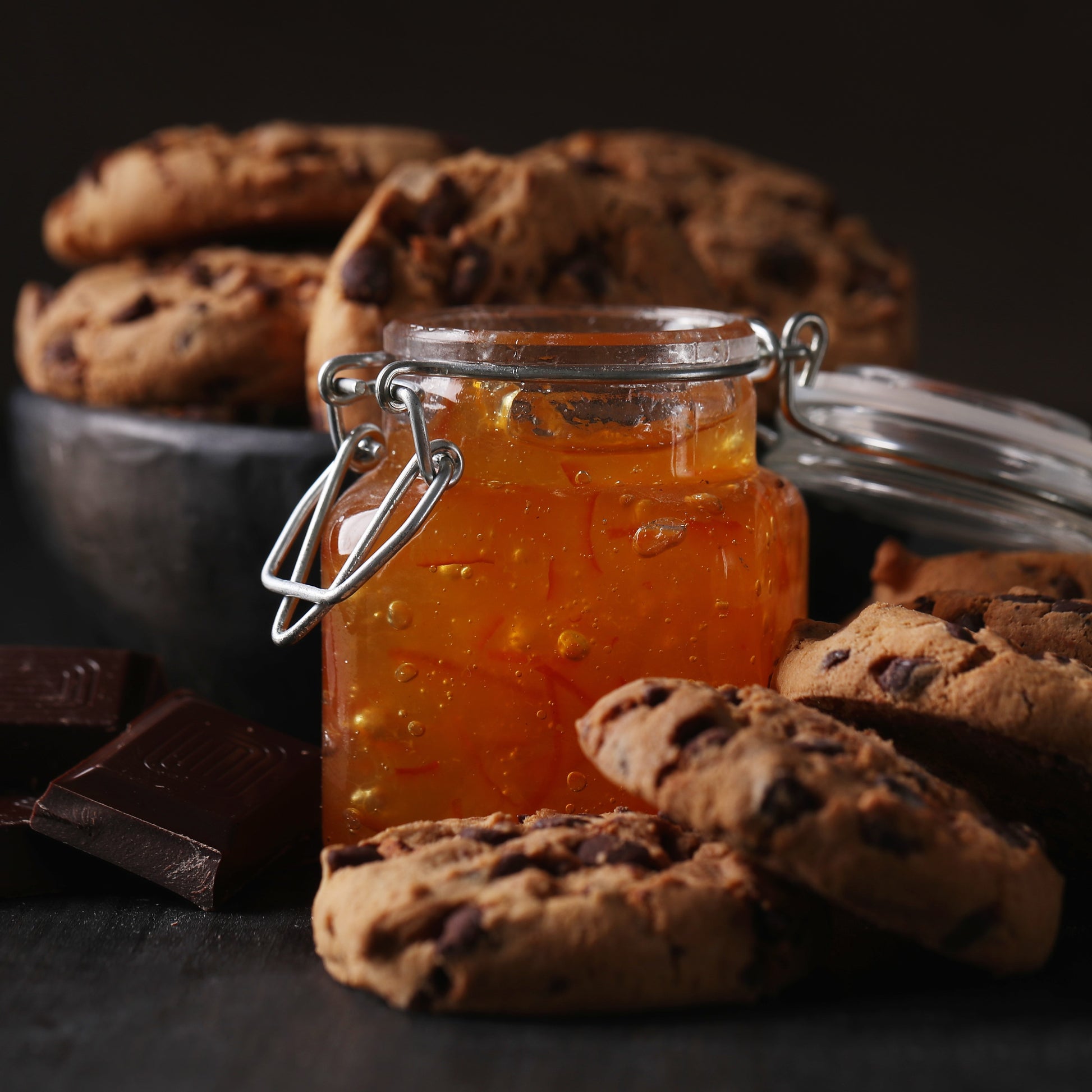Front view of several chocolate chip cookies placed around a square jar of apricot jam, with a bowl of more cookies and chunks of chocolate in the background.