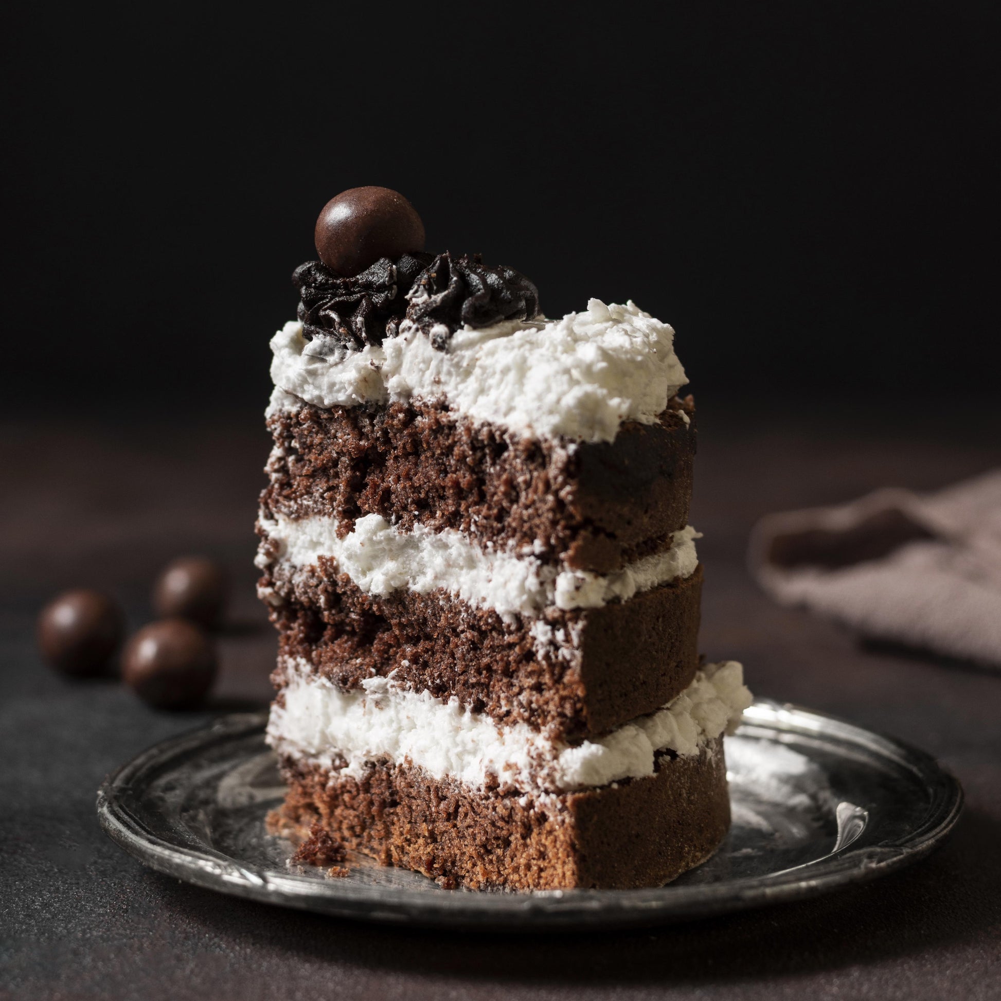 Front view of a moist chocolate cake slice on a porcelain plate, layered with cream cheese frosting, decorated with cocoa buttercream dollops, and topped with milk chocolate-covered coffee beans.