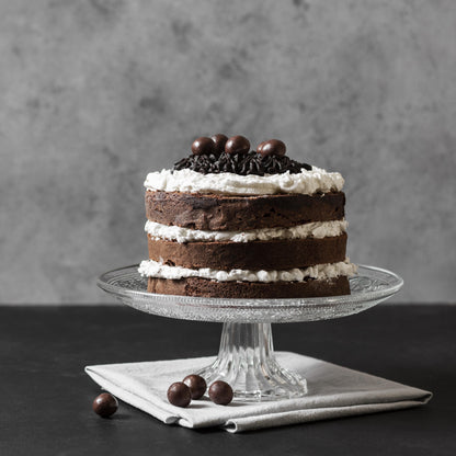 Front view of a moist chocolate cake on a glass cake stand, layered with cream cheese frosting, decorated with cocoa buttercream dollops, and topped with milk chocolate-covered coffee beans.