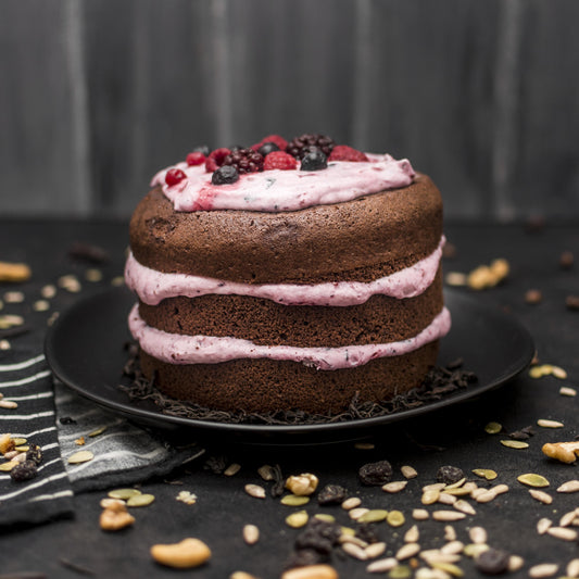 Front view of a cocoa pound cake with layered berries yogurt foam, and fresh berries, on a black plate with seeds scattered in the background.