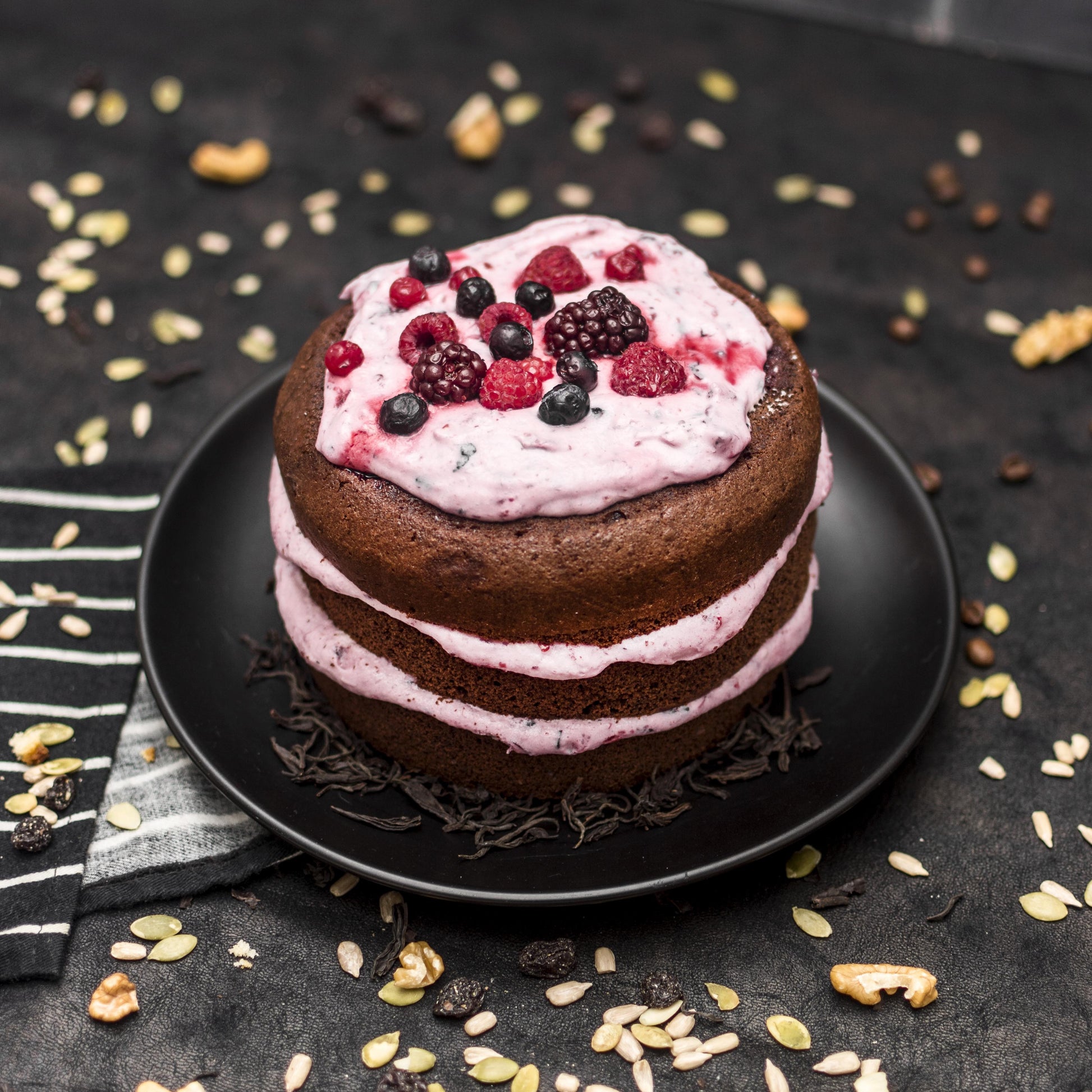 Angled top view of a cocoa pound cake with layered berries yogurt foam, and fresh berries, on a black plate with seeds scattered in the background.
