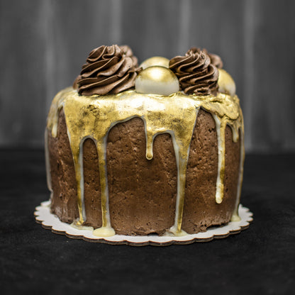 Front view of a salted caramel cream-filled sponge cake, covered with chocolate buttercream, drizzled with white chocolate ganache, edible liquid gold, topped with gold-dusted white chocolate caramel-filled balls, and chocolate buttercream swirls.