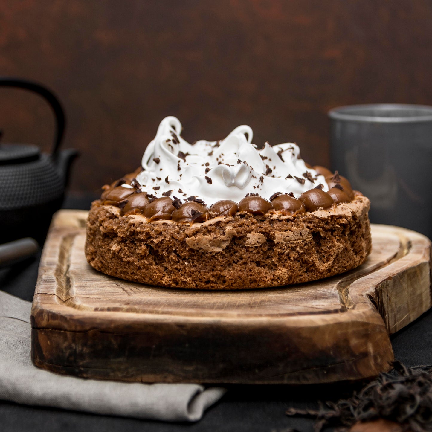 Front view of a brownie cake crowned with Italian meringue and salted caramel dollops, on a rustic wooden board with a cloth, mug and teapot in the background.