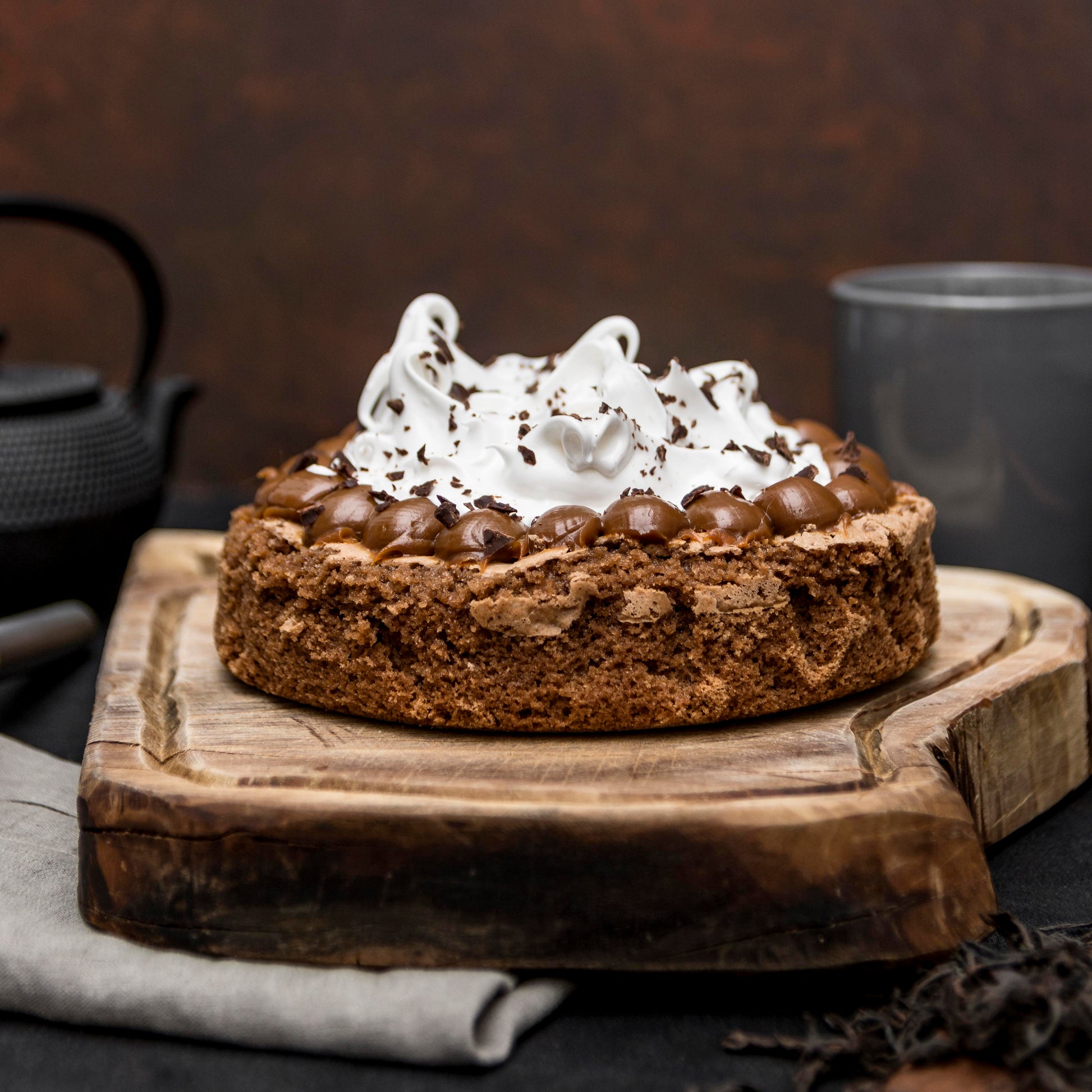 Front view of a brownie cake crowned with Italian meringue and salted caramel dollops, on a rustic wooden board with a cloth, mug and teapot in the background.