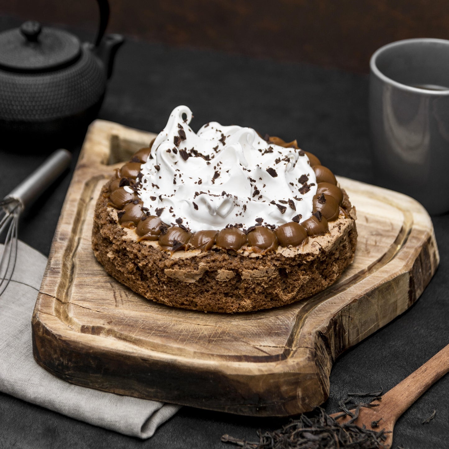 Angled top view of a brownie cake crowned with Italian meringue and salted caramel dollops, on a rustic wooden board with a cloth, mug, teapot, whisk, wooden spoon, and chocolate shavings in the background.