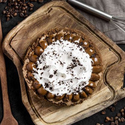 Top view of a brownie cake crowned with Italian meringue and salted caramel dollops, on a rustic wooden board with a cloth, mug with coffee, whisk, wooden spoon, and coffee beans in the background.